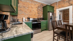 A kitchen with green cabinets, a brick wall, a stainless steel refrigerator, and a dining area with wooden chairs.