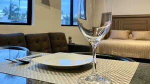 A hotel room with a bed, a glass table with a wine glass, a plate, and a view of palm trees through the window. - NEW! Ample Condo- Walk to Beach! AMAZING Location! Everything Walk-in distance! (Mazatlán)