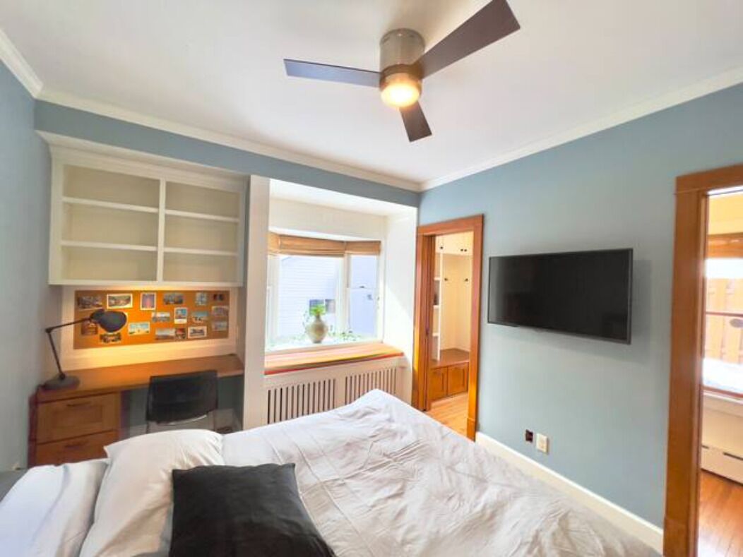 Ein Schlafzimmer mit einem Bett, einem Schreibtisch mit Stuhl, einem Deckenventilator, einem an der Wand befestigten Fernseher und einem Fenster mit Jalousien.