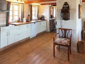 A kitchen with white cabinets, a wooden floor, a vintage clock, and a chair with a floral cushion.