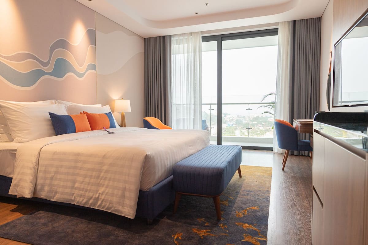 A modern hotel room with a large bed, a desk, and a balcony with a city view.