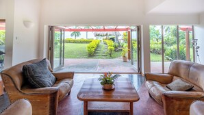 A spacious living area with leather sofas, a wooden coffee table, and a view of a lush garden through large sliding glass doors. - Malaqereqere Villas Ocean/Pool View, Villa 3 (Sigatoka)