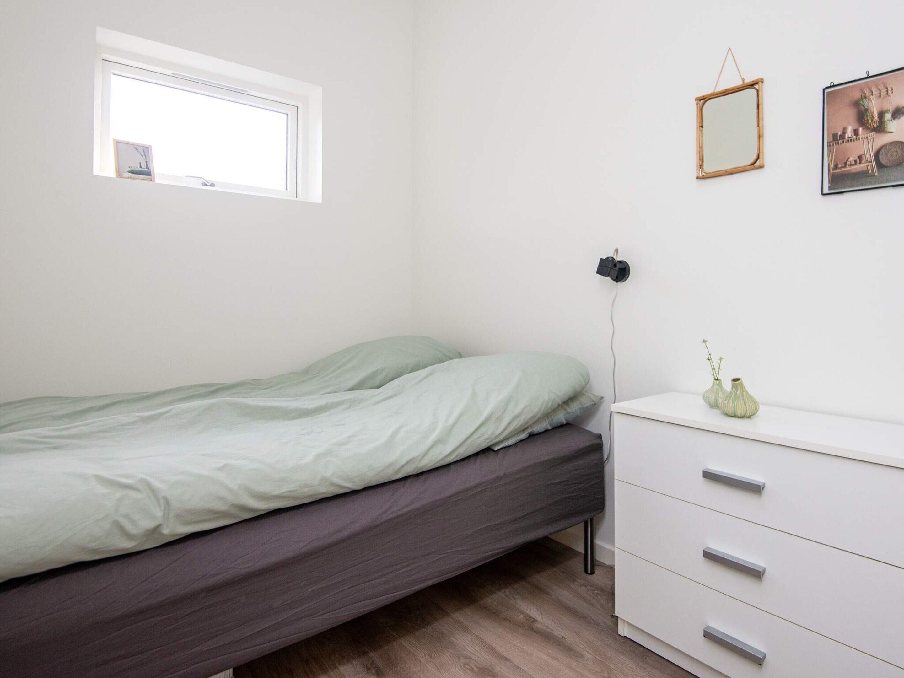 Un dormitorio minimalista con cama, mesita de noche, cómoda, ventana y espejo montado en la pared.