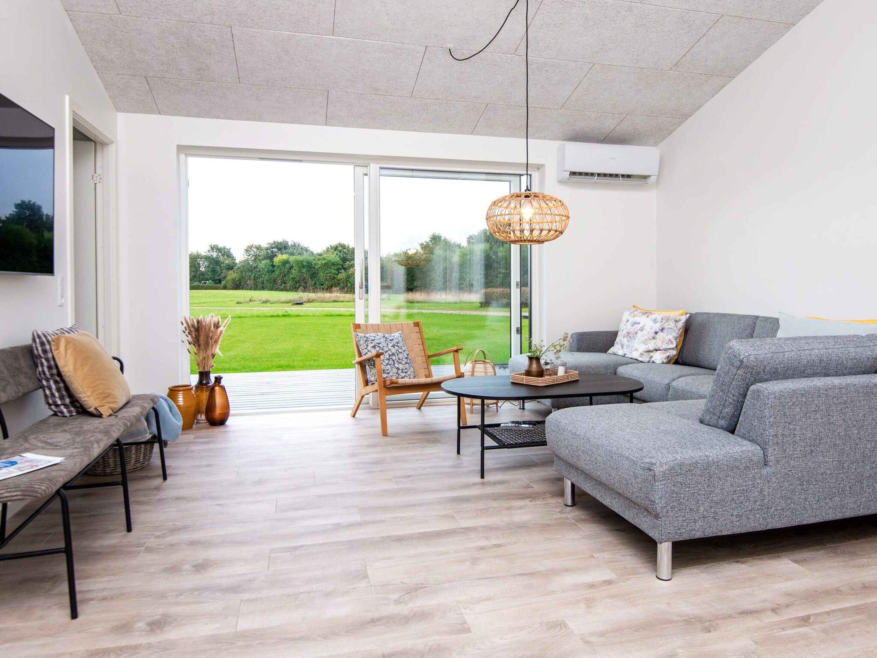 Un salon moderne avec un canapé gris, une chaise en bois, une table basse et une grande porte-fenêtre coulissante donnant sur un jardin.