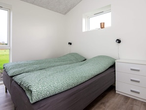 Ein Einzelbett mit einer grün gemusterten Tagesdecke, ein weißer Nachttisch mit Schubladen, ein kleines Fenster mit Blick ins Freie und eine Wandlampe.