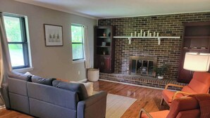 A living room with a brick fireplace, a grey sectional sofa, an orange armchair, and a wooden floor. - Country Lane Cottage, Privacy & Quiet Neighborhood (Rossville)