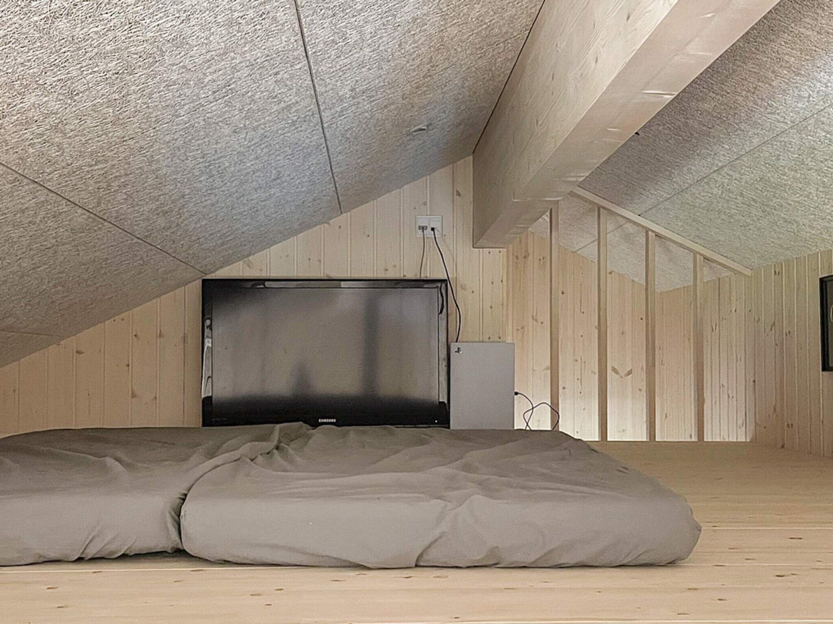 Una habitación con techo inclinado, un televisor de pantalla plana grande montado en la pared y una cama con colchón gris.