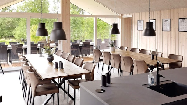 En rummelig spiseafdeling med lange træborde, moderne stole, store vinduer og hængende pendellamper.