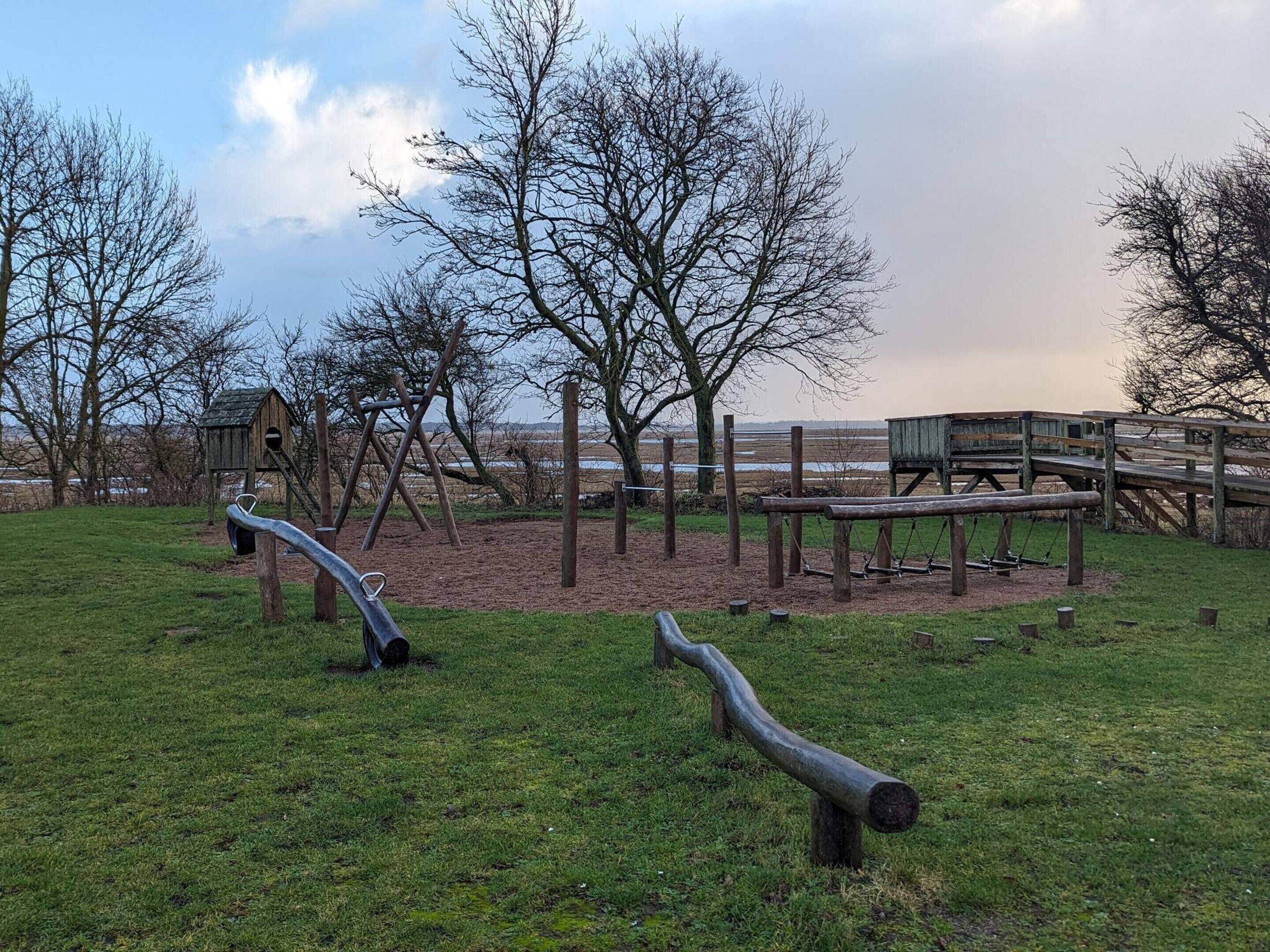 A playground with swings and a climbing structure, surrounded by trees and a wooden deck.
