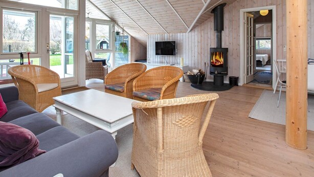 A cozy cabin interior with a large window, wicker furniture, a flat-screen TV, and a wood-burning stove. - 10 Person Holiday Home in Stege (Stege)