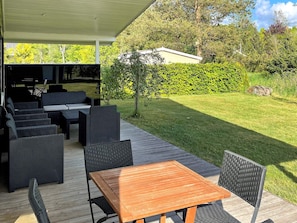 Ein möblierter Außenbereich mit Holztable und -stühlen, eine überdachte Lounge mit Sofas und Esstisch sowie ein gepflegter Rasen mit Hecke und Bäumen im Hintergrund.