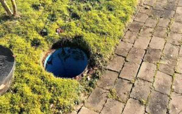 A circular hole in the ground surrounded by grass, next to a paved walkway.