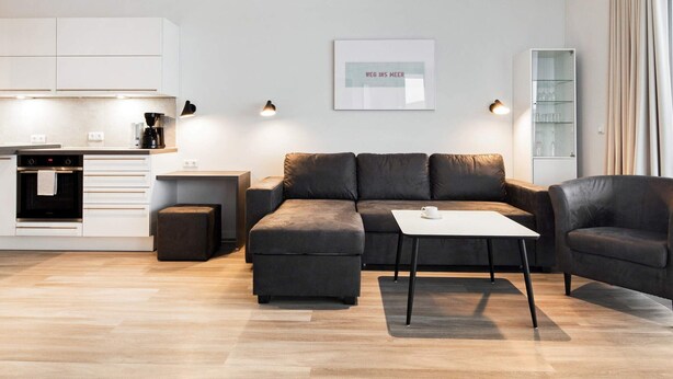A modern living room with a sofa, ottoman, and coffee table, a kitchen area with cabinets and appliances, and a framed artwork on the wall.
