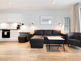 En moderne stue med sofa, skærmstole og sofabord, et køkkenområde med skabe og hårde hvidevarer, samt et billede i ramme på væggen.