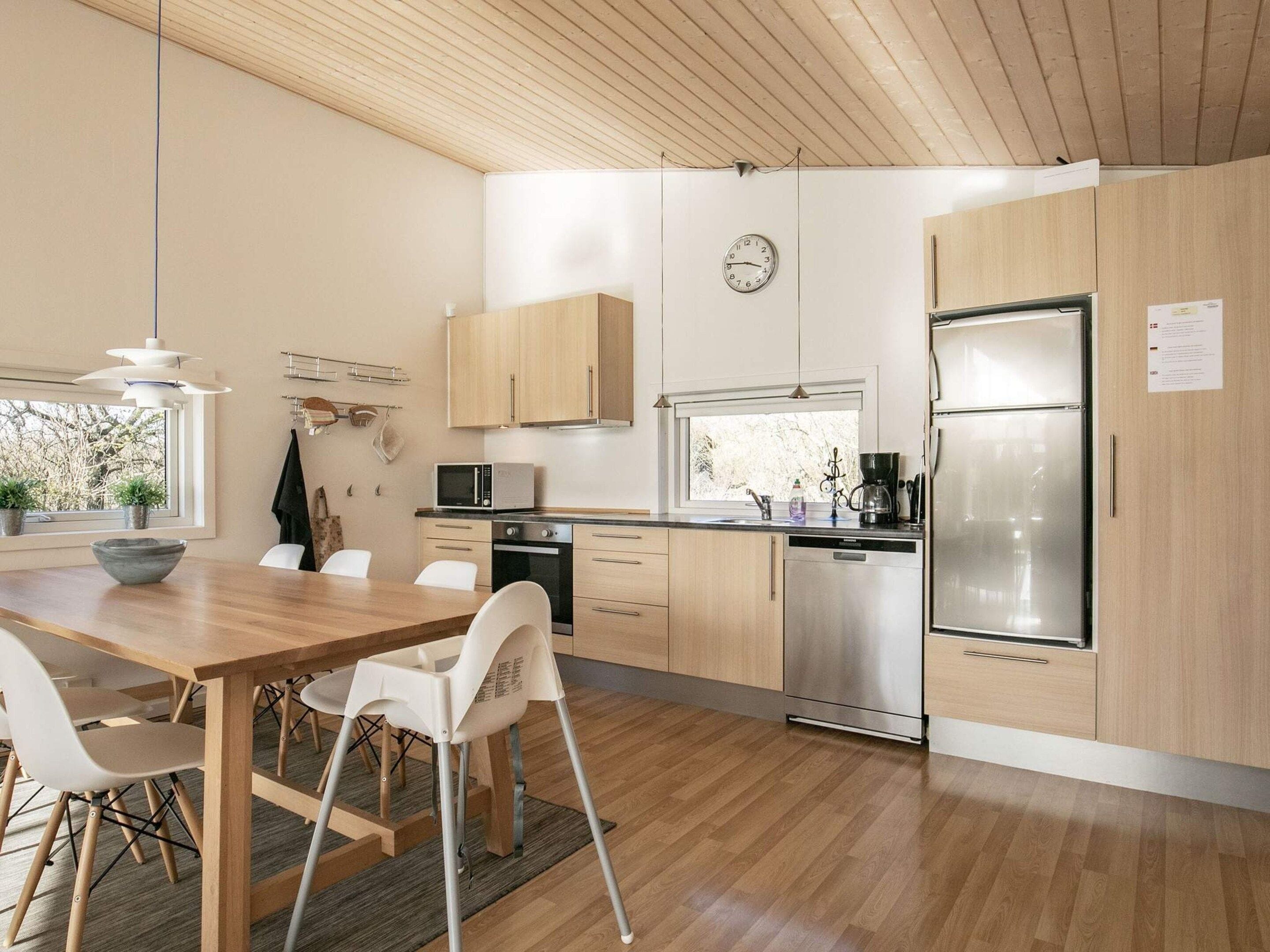 A modern kitchen with wooden cabinets, a stainless steel refrigerator, a microwave, and a dishwasher. There is a dining table with chairs and a window providing natural light.