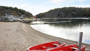 A beach with two red boats tied to a wooden post, a sandy shore, and a calm lake with a dock and buildings in the background. - 5 Person Holiday Home in Stromstad (Stromstad)