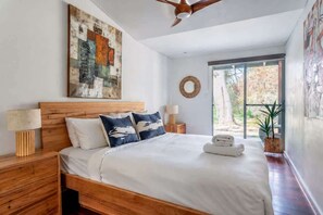 A bedroom with a wooden bed, a ceiling fan, a large abstract painting, a round mirror, and a view of the outdoors through a sliding door.