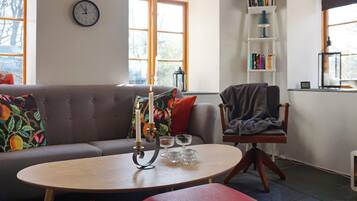 A cozy living room with a sofa, a round table with candles, a bookshelf, and a clock on the wall.