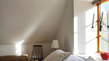 A bedroom with a sloped ceiling, a bed with white bedding, a wooden dresser, a chair, a lamp, and a window with a curtain.
