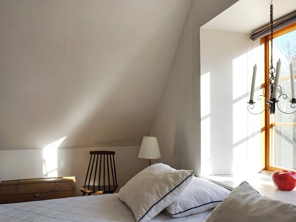 Un dormitorio con techo inclinado, una cama con ropa de cama blanca, un aparador de madera, una silla, una lámpara y una ventana con cortina.