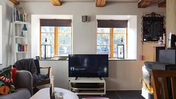 A cozy living room with a television, bookshelves, a red ottoman, and wooden beams on the ceiling.