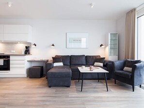 Un salon moderne avec un canapé, un pouf et une table basse. On y trouve également un coin cuisine avec des placards et une machine à café. Un tableau encadré est accroché au mur, et il y a une fenêtre avec des rideaux.
