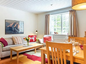Una sala de estar acogedora con un sofá, una mesa de centro y una mesa de comedor para dos. Se ve una ventana con cortinas y vistas a la vegetación.