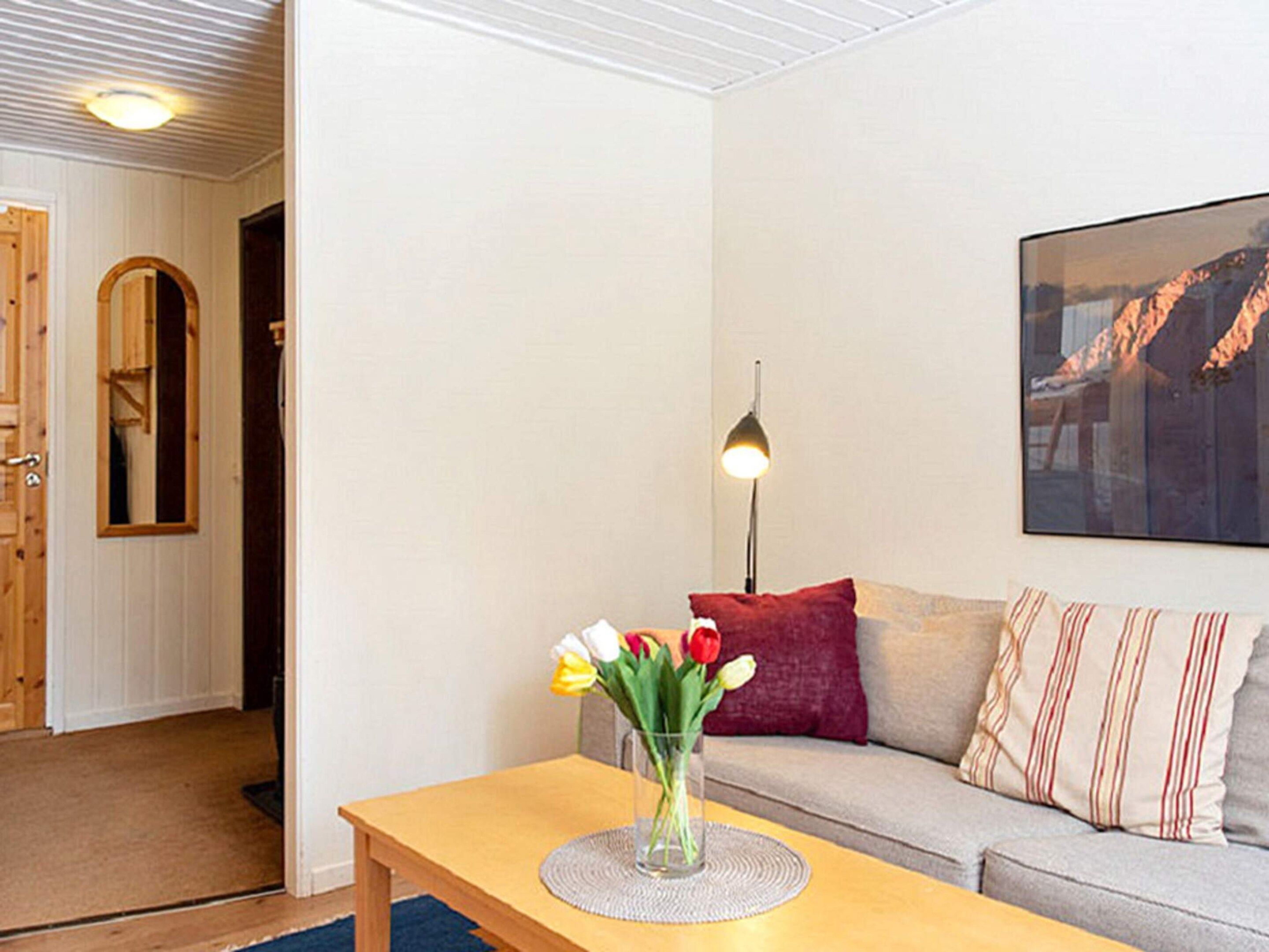 A living room with a wooden table, a vase with flowers, a sofa with cushions, a lamp, and a framed picture on the wall.
