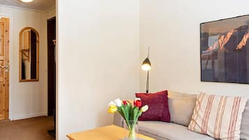 A living room with a wooden table, a vase with flowers, a sofa with cushions, a lamp, and a framed picture on the wall.