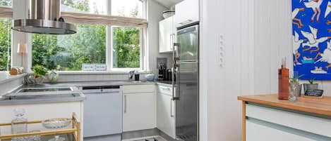 A modern kitchen with a stainless steel refrigerator, a dishwasher, and a window with a view of greenery.