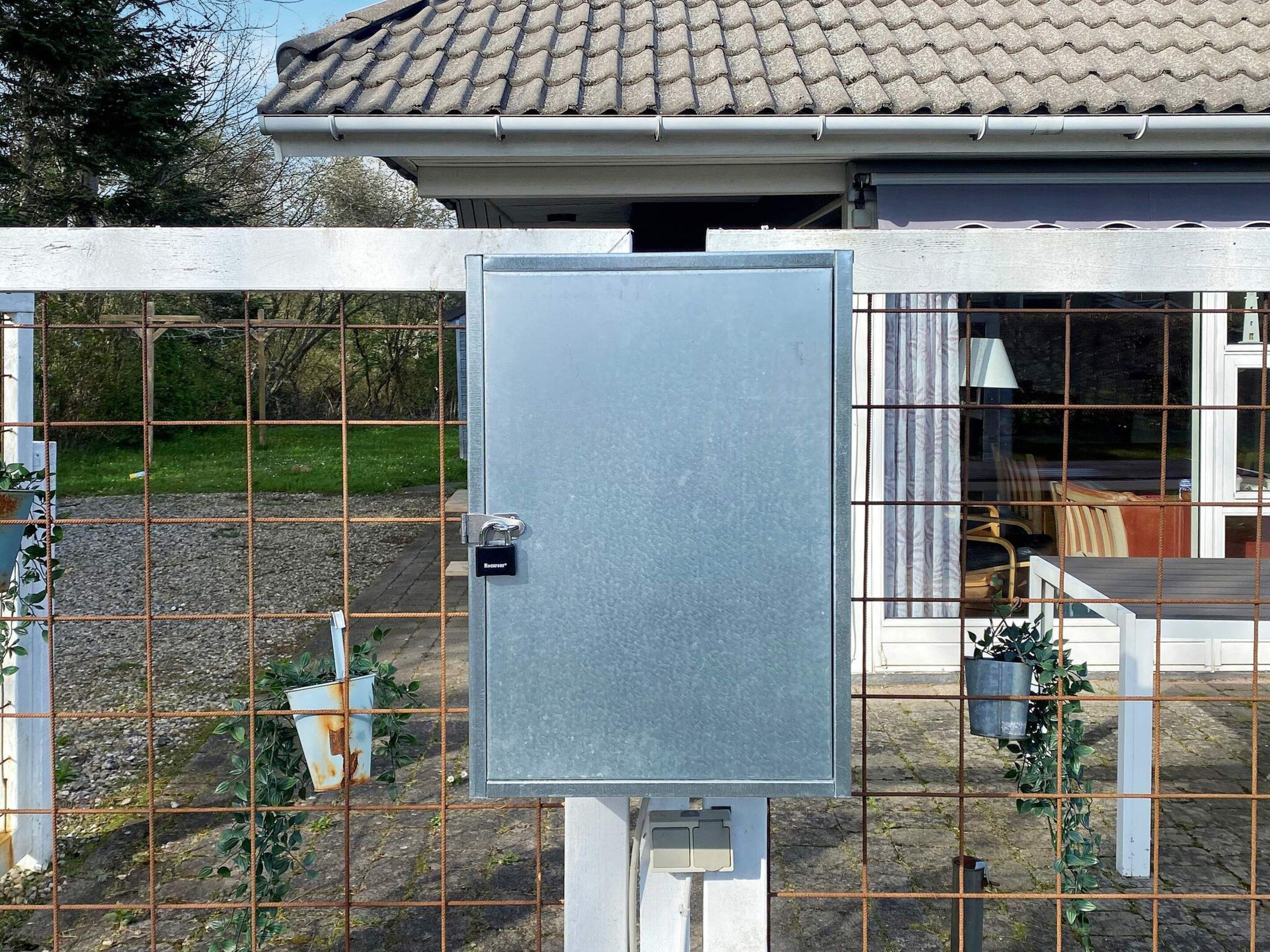 Une boîte électrique en métal, fermée à clé à l’aide d’un cadenas, entourée d’une clôture métallique et agrémentée de plantes.