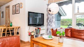 Un accogliente soggiorno con soffitto in legno, una TV a schermo piatto, una libreria, un divano in pelle e un'ampia finestra vista su un edificio e vegetazione.