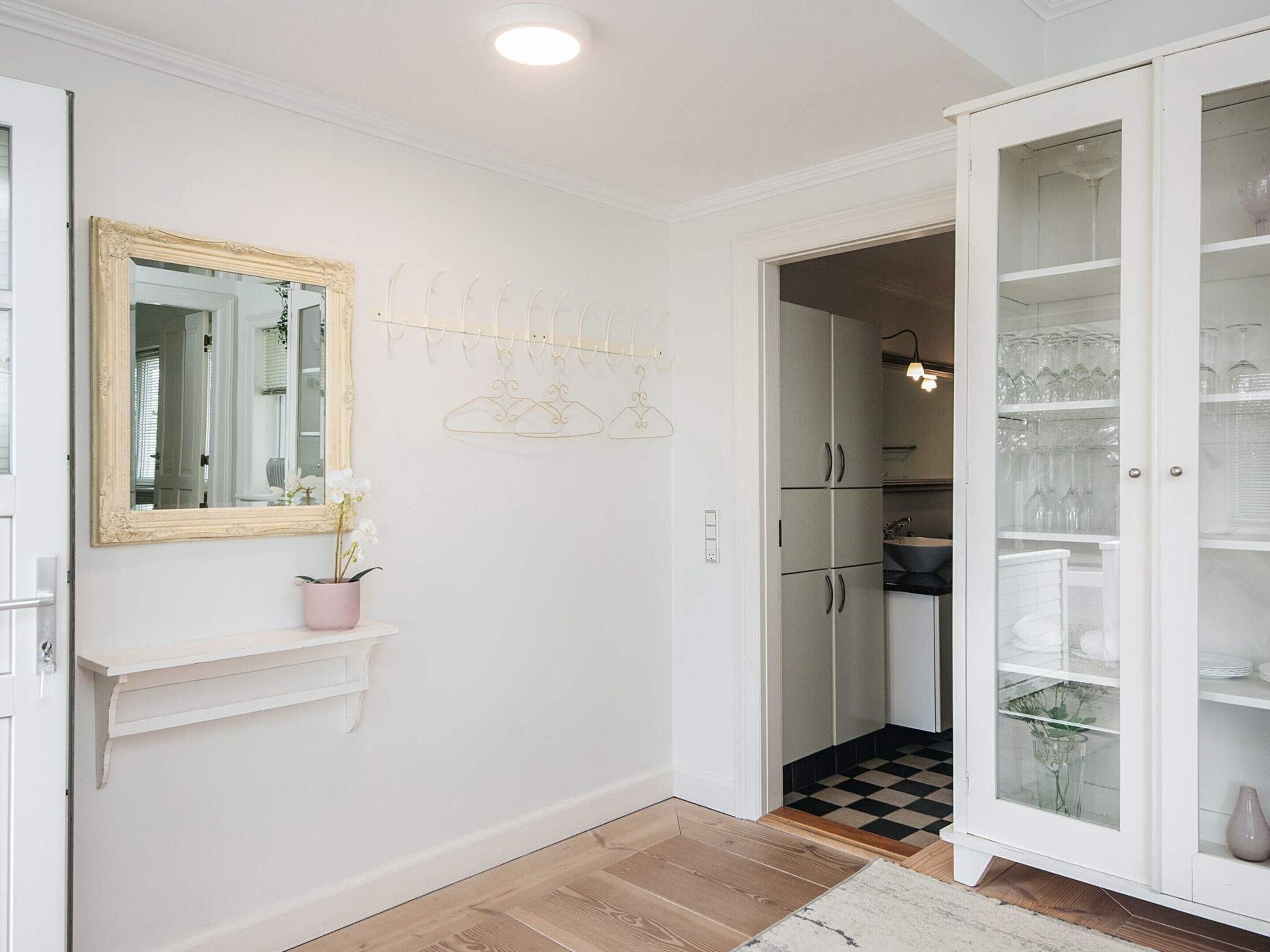 A hallway with a white door, a mirror with an ornate frame, a pink vase with flowers, a coat rack, and a cabinet with glass doors.