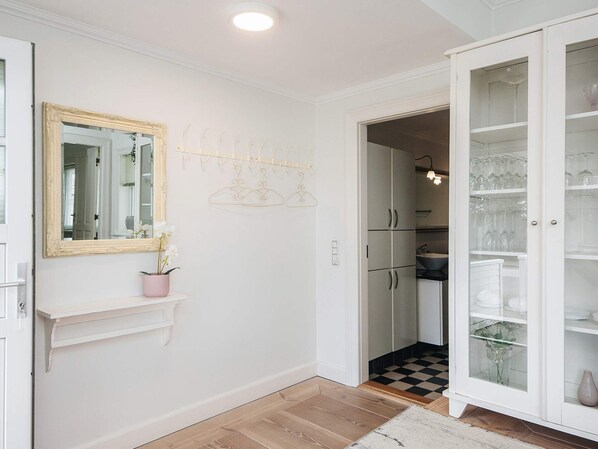 A hallway with a white door, a mirror with an ornate frame, a pink vase with flowers, a coat rack, and a cabinet with glass doors.