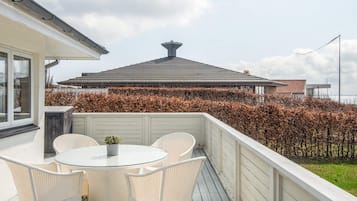 A balcony with a round glass table and wicker chairs, overlooking a building with a sloped roof and a hedge in the background.