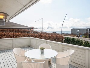 En overdækket udendørs siddeplads med et rundt bord og stole, med udsigt over kysten med en bygning og en vej.