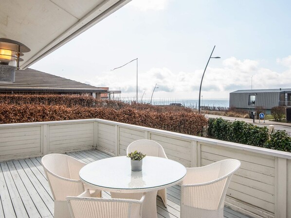 A covered outdoor seating area with a round table and chairs, overlooking a coastal view with a building and a road. - 5 Star Holiday Home in Esbjerg V (Esbjerg)