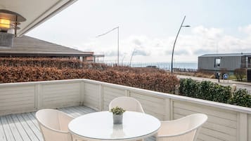 A covered outdoor area with a round glass table and wicker chairs, overlooking a body of water and a distant building.