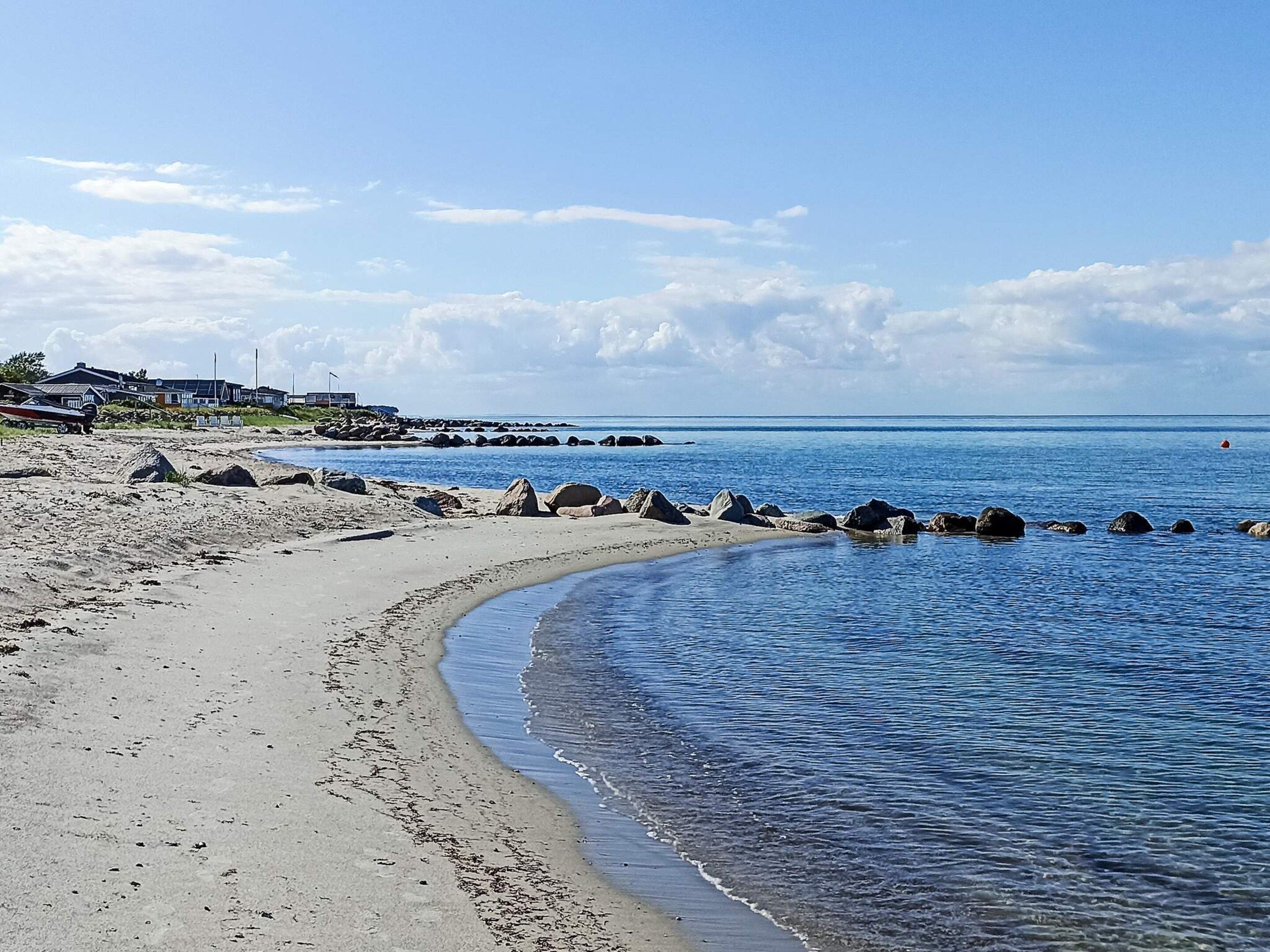 Una playa con una orilla curva, un rompeolas rocoso y casas al fondo.