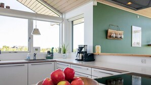 A modern kitchen with a wooden bowl of apples on the counter, a coffee maker, and a view of the outdoors through large windows. - 9 Person Holiday Home in Otterup-by Traum (Otterup)