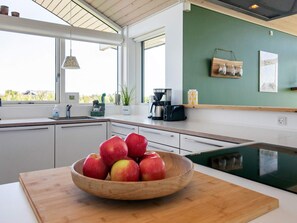 A modern kitchen with a wooden bowl of apples on the counter, a coffee maker, and a view of the outdoors through large windows. - 4 Star Holiday Home in Otterup (Otterup)
