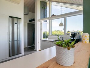 Et moderne k økken med et rustfrit stål køleskab, en hvid bordplade og et stort vindue med udsigt til grøn vegetation udenfor.