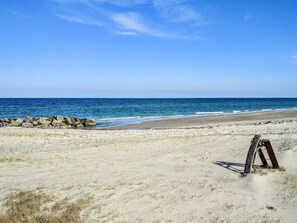 En strand med en klippefyldt bølgebryder, klar blå himmel og roligt hav.