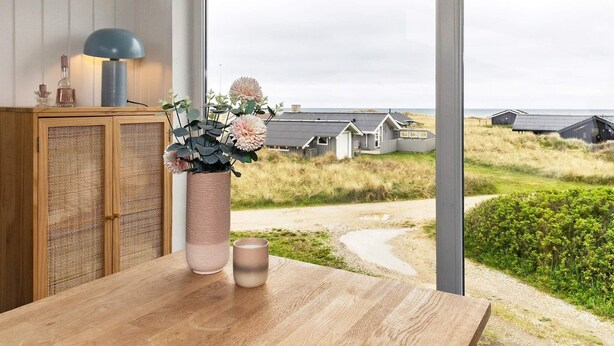 A wooden table with a vase of flowers and a mug, a cabinet with a blue lamp, and a view of houses and beach through large windows. - 8 Person Holiday Home in Hjorring (Hjørring)