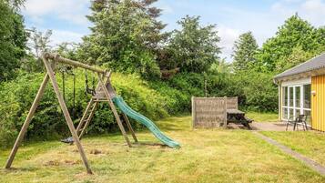 A wooden swing set with a green slide in a grassy backyard with a wooden fence and a yellow building.