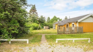 A gravel path leading to a wooden bench, a swing set, and a yellow building with a fenced yard.