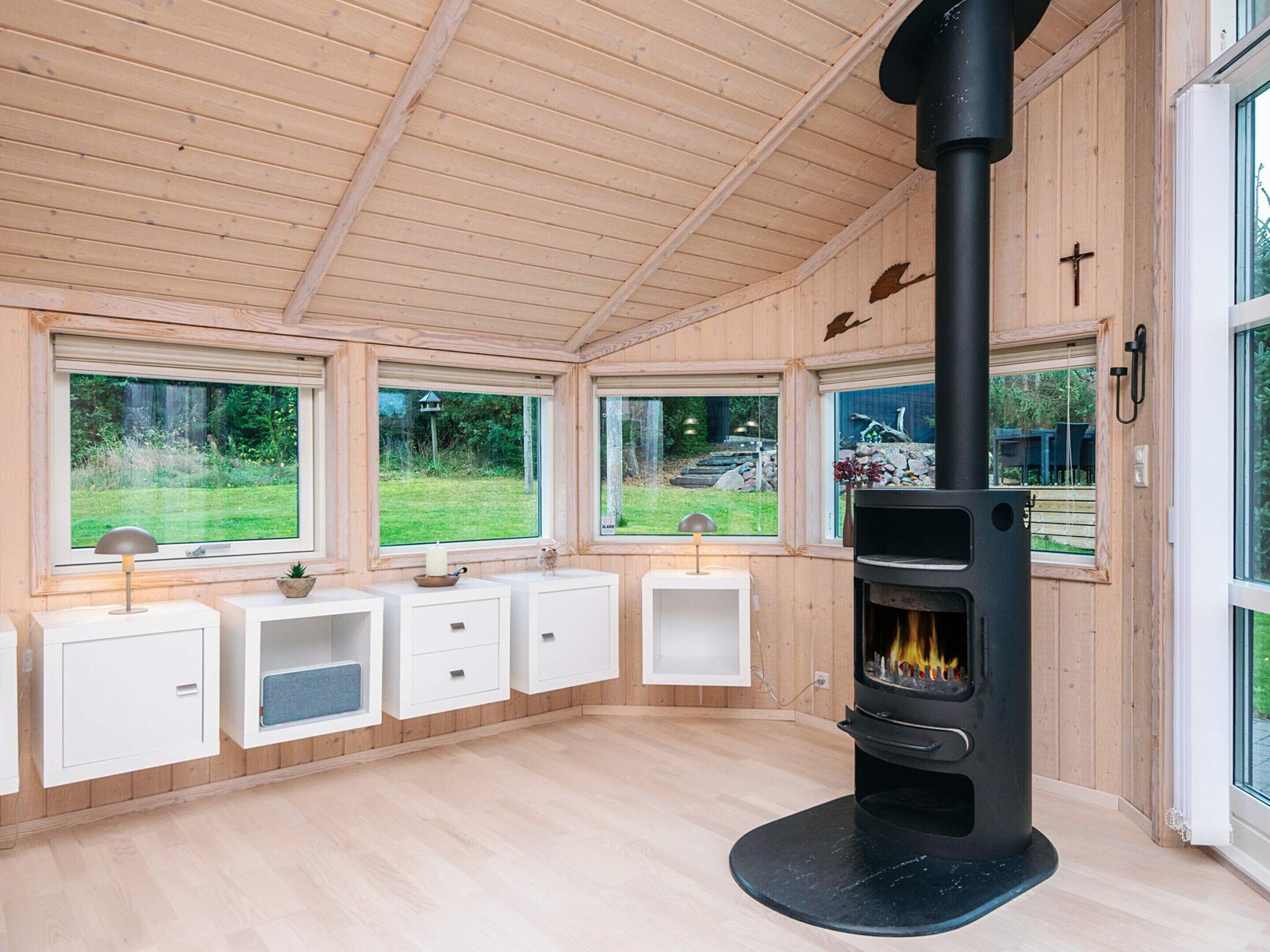 A wooden interior with a central black stove, white cabinets, and large windows offering views of a garden.