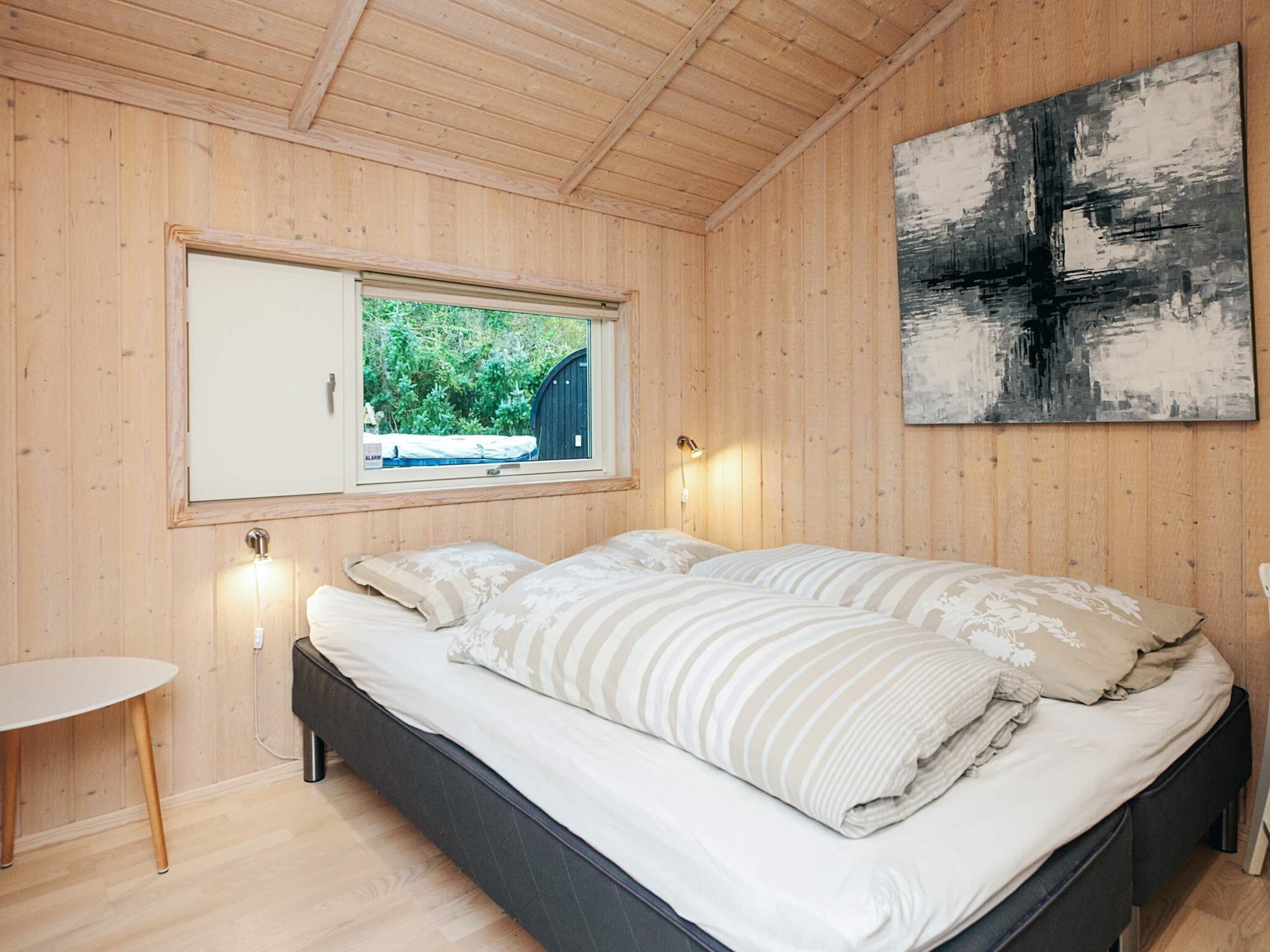 A bedroom with a bed, a window, a wall-mounted artwork, and a small table.