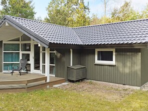 A small, single-story building with a grey tiled roof, a covered porch with a chair, and a window. - 6 Person Holiday Home in Frederiksvaerk (Frederiksvaerk)