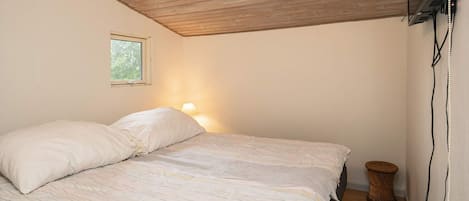 Une chambre avec un plafond en bois, un lit avec une literie blanche, une lampe de chevet, une petite fenêtre et un téléviseur fixé au mur.